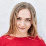 Mladá žena s klidným výrazem, dlouhými blond vlasy a modrýma očima se jemně usmívá. Na sobě má červený top a je na bílém pozadí.