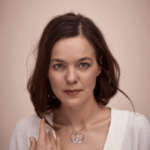 Woman with serene expression, dark hair, wearing a white V-neck top and a silver pendant. Her hand lightly touches her collar
