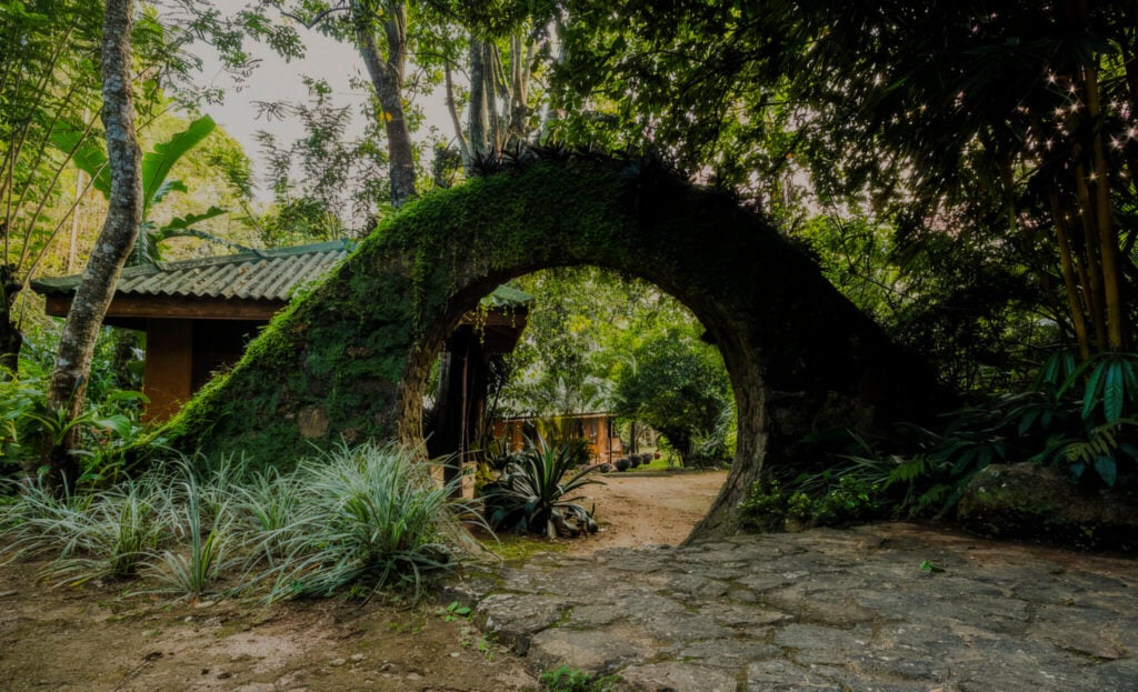 Vstupní brána z kamene porostlého mechem do luxusního wellness resortu, ukrytého v srdci bujného srílanského mlžného pralesa.