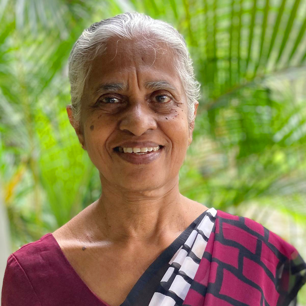 Elderly South Asian woman, possibly an Ayurvedic practitioner, smiling gently. She wears traditional attire with a green, lea