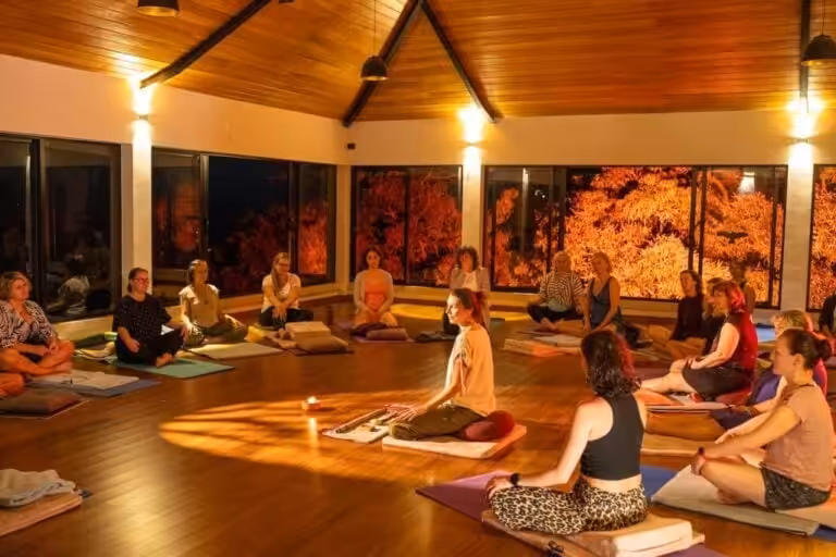 A group of people sitting in a circle on yoga mats during a guided meditation session in a warmly lit, wood-paneled studio.