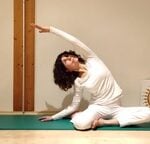 A woman in white clothing performs a seated side-bend yoga pose on a green mat in a softly lit room with a candle.