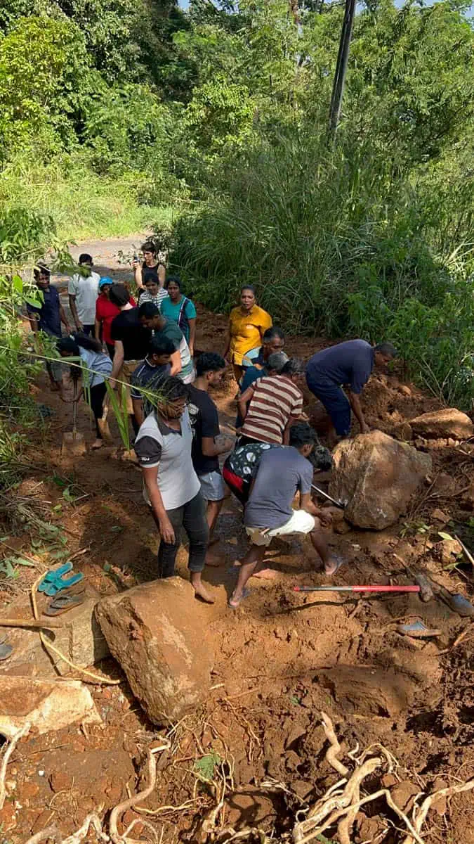 A group of people work together to clear mud and large rocks from a rural road after a landslide.