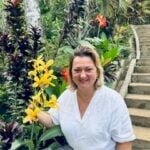A woman smiles while touching a stalk of bright yellow flowers in a lush tropical garden with stone steps.