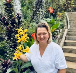 A woman smiles while touching a stalk of bright yellow flowers in a lush tropical garden with stone steps.