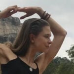 A woman in a black top holding a serene yoga pose, with her arms raised above her head against a backdrop of rock formations