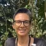A smiling Yasmin Hill guest with short hair and glasses, set against a lush green background.