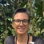 A smiling Yasmin Hill guest with short hair and glasses, set against a lush green background.