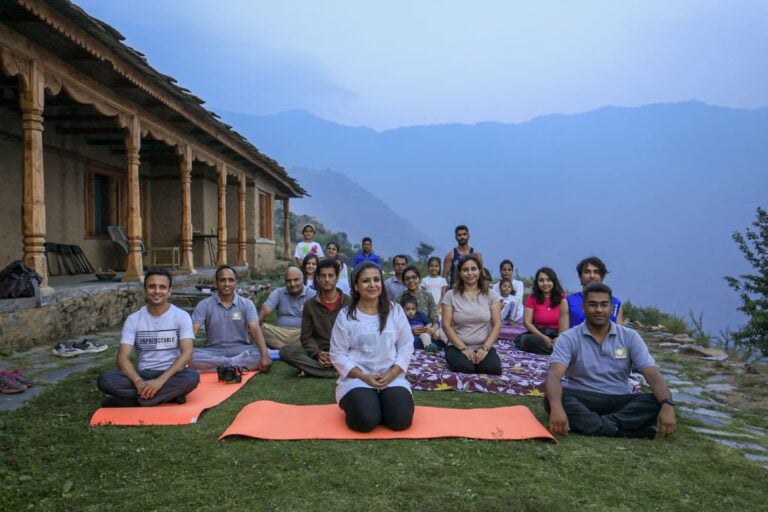 Bhawna Chhabra with a retreat group seated on yoga mats in front of a wooden mountain house at dusk.